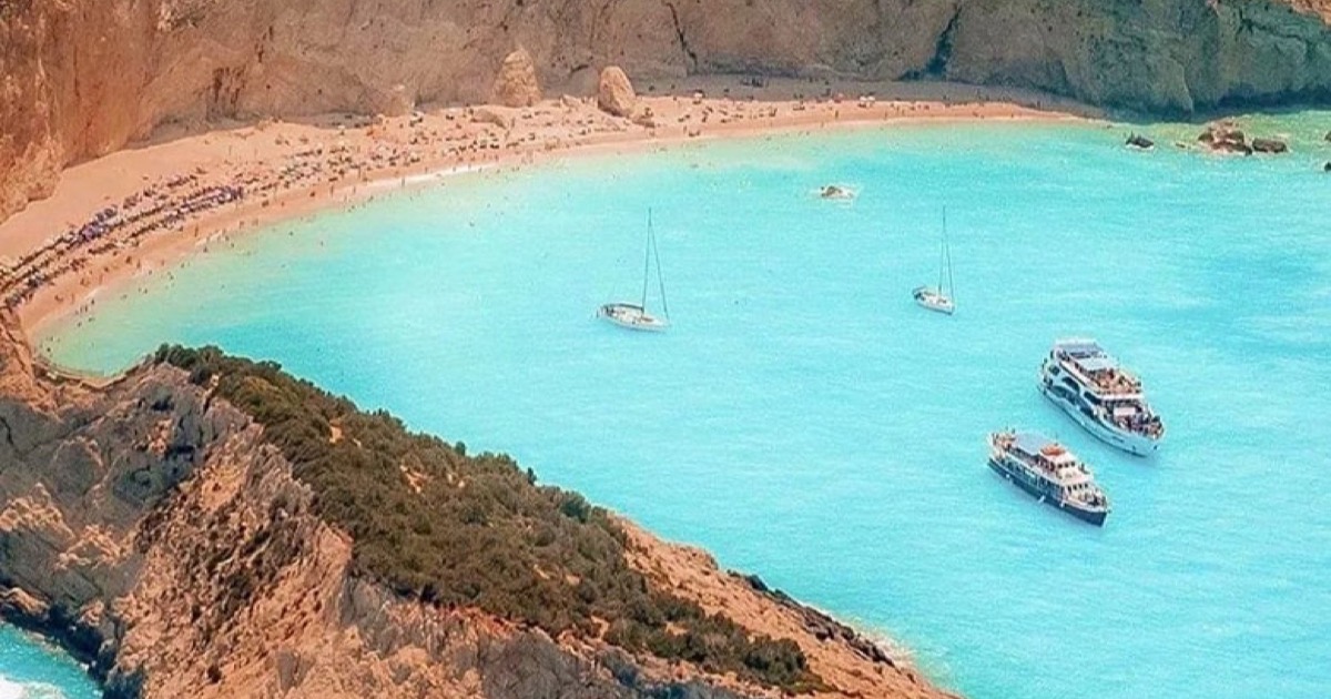 Yachts anchored off a sandy Greek beach below red cliffs, seen from the air.