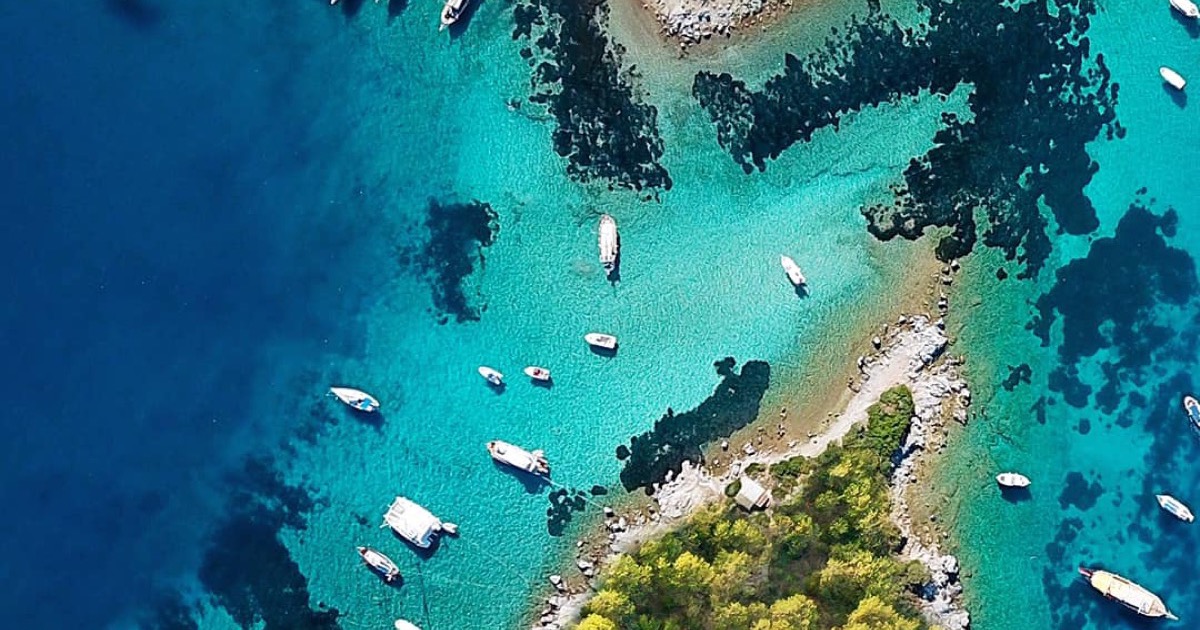 Boats anchored over turquoise shallows along a rocky Turkish shoreline, seen from the air.